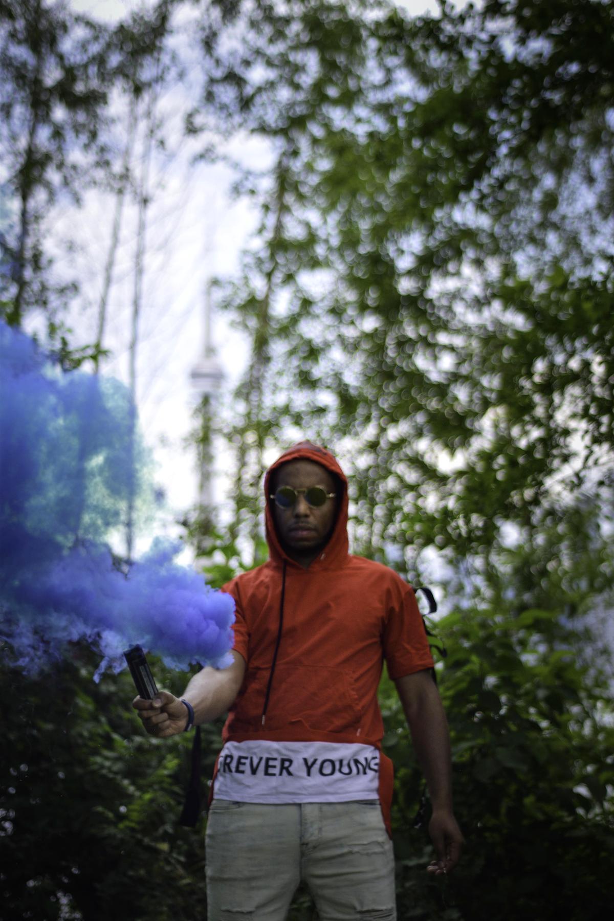 NoMatch in a red hoodie writing and performing outdoors with blue smoke in the background